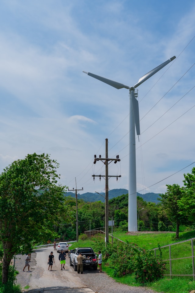 People walk near a tall wind turbine under a cloudy sky.
